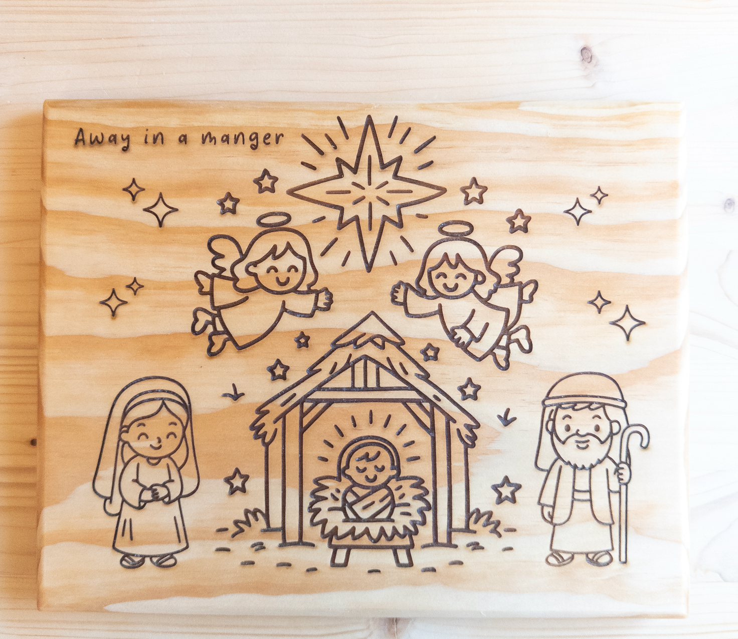 Wooden plaque with engraved nativity scene and text 'Away in a manger' on a wooden background.